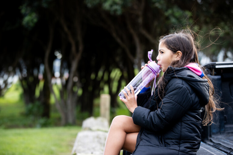 Young girl drinking from purple water bottle while sitting outdoors in park setting